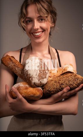 Panificia sorridente che tiene una varietà di pani artigianali freschi, offrendo prodotti da forno fatti in casa Foto Stock
