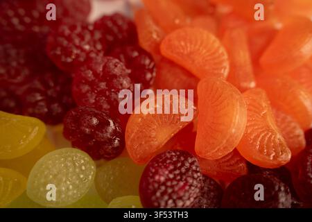 Macro primo piano di caramelle gommose lucide al gusto di frutta ammucchiate su un tavolo: fette di mandarini arancioni traslucide, gommini rosso scuro e dischi giallo-verdi Foto Stock