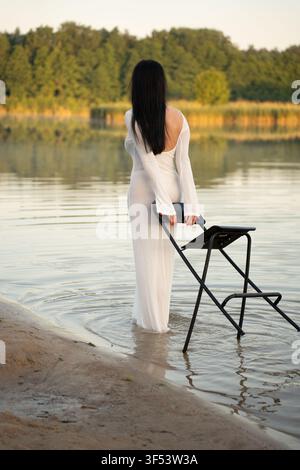 Donna in un vestito bianco traslucido in piedi sulla riva del lago durante l'ora dorata, che tiene una sedia nera piegata e guarda riflessi fiancheggiati da alberi sul cal Foto Stock