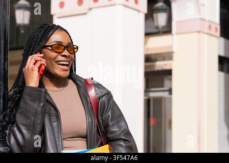 Giovane donna nera con trecce e occhiali da sole che tiene libri e parla felicemente su smartphone in una strada cittadina Foto Stock