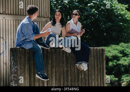 Tre studenti seduti su un muro di cemento all'aperto, chiacchierando e studiando insieme nel campus Foto Stock