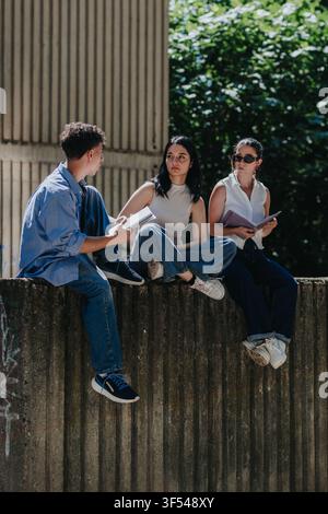 Tre studenti seduti su un muro di cemento che parlano e studiano insieme all'aperto nel campus Foto Stock