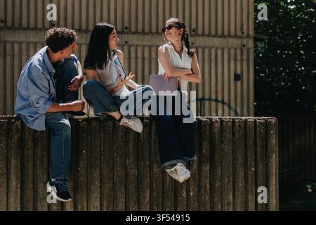 Tre studenti che chiacchierano su un muro del campus durante una pausa studio informale all'aperto Foto Stock