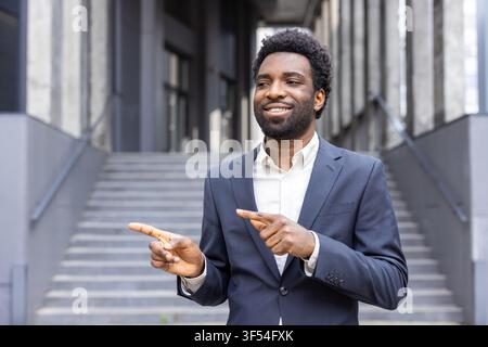 Uomo africano in abiti formali che sorride e indica con entrambe le mani a sinistra e a destra, invitando gli spettatori a esplorare diverse opportunità o direzioni ou Foto Stock