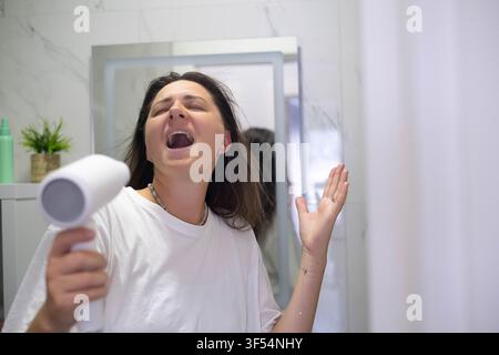Giovane donna in pigiama che si diverte asciugandosi i capelli, cantando e ballando in bagno, mostrando una routine quotidiana spensierata e gioiosa. Foto Stock