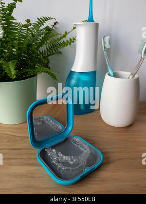 Una custodia blu aperta per retainers dentali con uno specchio e allineatori trasparenti posizionati su un tavolo di legno accanto a un irrigatore dentale, spazzolini in un portaspazzolino, un Foto Stock