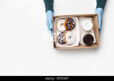 Lavoro di un ristorante fast food durante un'epidemia virale. Il cameriere consegna una scatola di ciambelle glassate al bianco e al cioccolato con briciole e due tazze da asporto Foto Stock
