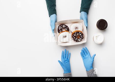 Tortine per il cliente a casa e problema con l'ordine. Cliente con guanti di gomma prende una scatola con ciambelle con glassa al cioccolato e bianca con briciole e due tazze di Foto Stock