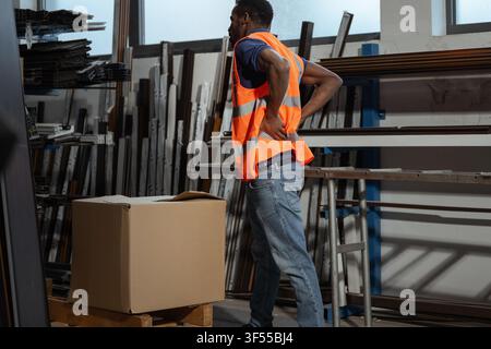 Un uomo in un magazzino di fabbrica sente dolore nella parte bassa della schiena mentre solleva una scatola di cartone pesante. Foto Stock