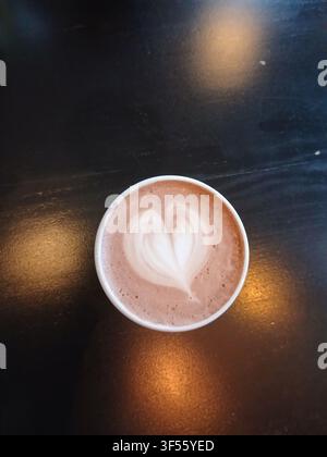 Vista dall'alto di una tazza di ceramica riempita di cioccolata calda con un motivo a cuore di schiuma di latte su un tavolo di legno nero Foto Stock