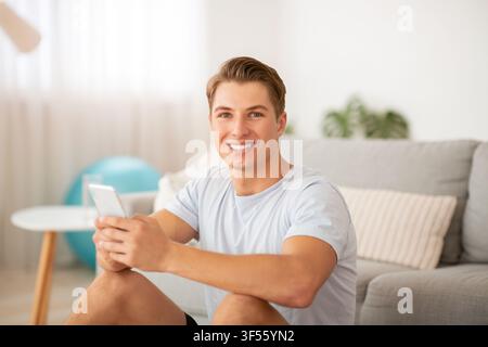Sorridente giovane che tiene smartphone, seduto all'interno del soggiorno con palla da fitness Foto Stock