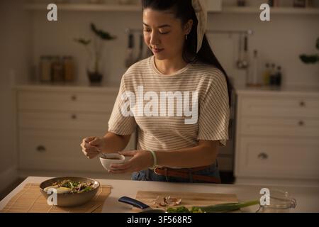 Donna asiatica che mescola la salsa per insalata in una piccola ciotola bianca con un cucchiaio di metallo al bancone della cucina Foto Stock
