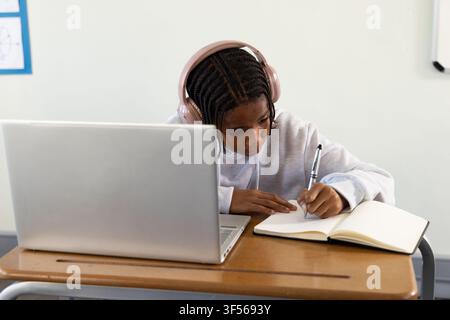Adolescente afroamericano che scrive appunti usando un laptop alla scrivania della scuola, indossando un auricolare rosa e una felpa con cappuccio Foto Stock