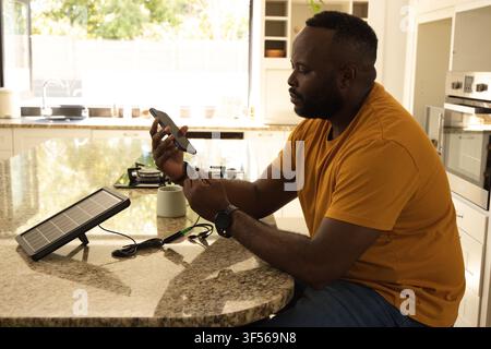 Uomo afroamericano che testa un cavo telefonico sull'isola della cucina vicino a un pannello solare, spazio di copia Foto Stock