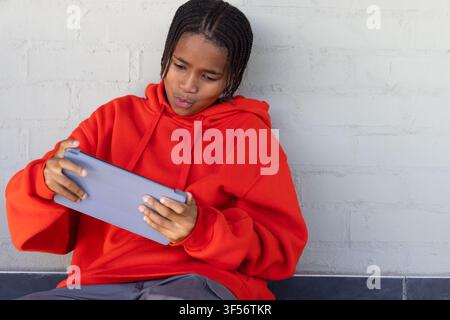 Adolescente afroamericano con felpa con cappuccio rossa seduto contro un muro grigio su un bordo scuro tenendo un tablet grigio Foto Stock