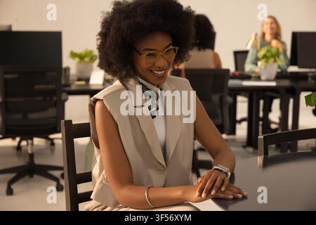 Donna afroamericana sorridente e seduta a un tavolo in ufficio, indossa un gilet beige e uno smartwatch Foto Stock