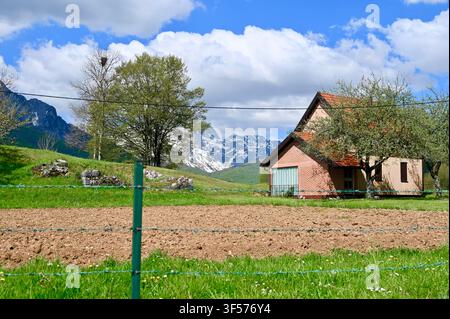 Un paesaggio rurale pittoresco con una piccola casa di legno, un campo arato in primo piano e maestose montagne innevate sotto un cielo luminoso e nuvoloso. Foto Stock