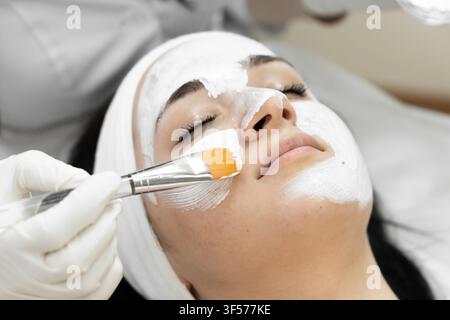 Primo piano di una giovane donna che si gode una maschera rigenerante in un salone di bellezza. L'estetista applica con cura il prodotto per la cura della pelle, sottolineando l'autocura e una pelle sana Foto Stock