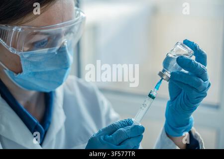 Prima vista di un medico che indossa mascherina, guanti e occhiali protettivi mentre preleva il vaccino da una fiala in una siringa. L'immagine evidenzia l'assistenza sanitaria moderna, l'immunizzazione e la prevenzione delle infezioni in un ambiente medico Foto Stock