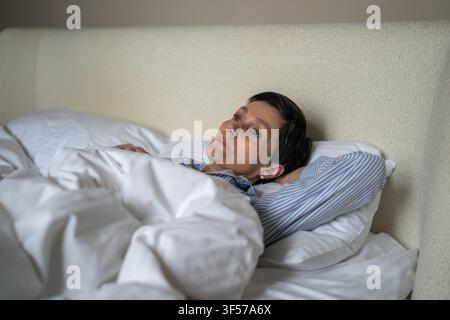 Infelice e premurosa donna anziana sdraiata a letto la mattina a casa con un'espressione persa e esausta. Foto Stock