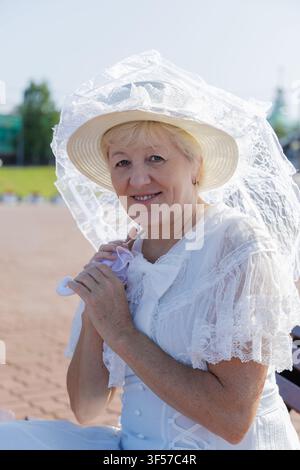 Donna matura sorridente in un abito vintage di pizzo bianco seduta su una panchina moderna in legno con un libro e un ombrellino. Foto Stock