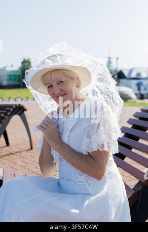 Donna matura sorridente in un abito vintage di pizzo bianco, seduta su una panca moderna di legno con un libro e un ombrellino. Foto Stock