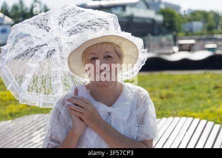 Donna matura sorridente in un abito vintage di pizzo bianco e cappello, che tiene un bellissimo parasole di pizzo abbinato in un parco soleggiato. Foto Stock