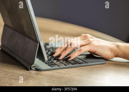 Primo piano di mani sottili di donna che digitano su una tastiera per tablet a una scrivania di legno. Spazio di lavoro mobile moderno, dispositivo digitale, lavoro da remoto, comunicazione online, libero Foto Stock
