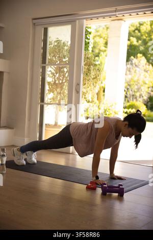 Donna africana adulta che esegue un plank alto su un tappeto scuro a casa con manubri e una bottiglia d'acqua Foto Stock