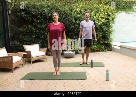 Coppia matura che pratica esercizi su materassini da yoga verdi, bottiglie d'acqua sul patio, indossa abbigliamento sportivo Foto Stock