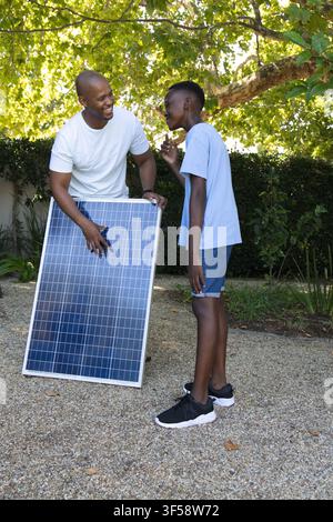 Padre afroamericano e figlio adolescente che tengono ed esaminano un grande pannello solare nel giardino sul retro Foto Stock