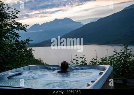 Valdez, Alaska. Una donna si rilassa in una vasca idromassaggio la sera con vista sul lago Robe durante una vacanza in Alaska. Foto Stock