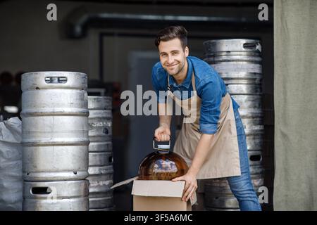 Giovane uomo caucasico sorridente che solleva un grande barattolo di vetro in una birreria, concetto di produzione di alcol Foto Stock