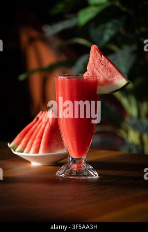 Succo di anguria fresco, una fetta di anguria come guarnizione, accanto a un piatto di fette di anguria su un tavolo di legno con piante verdi sullo sfondo. Foto Stock