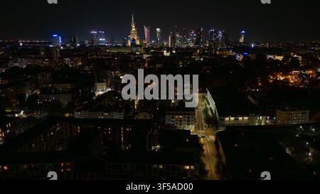 Veduta aerea notturna del centro di Varsavia che mostra grattacieli luminosi, una torre monumentale con cupola, blocchi residenziali fitti, edifici per uffici, finestre luminose e tracce di traffico sulle strade trafficate Foto Stock
