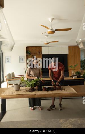 Coppia di anziani diversificati che piantano piante a un lungo tavolo di legno nell'area giorno con carta kraft Foto Stock