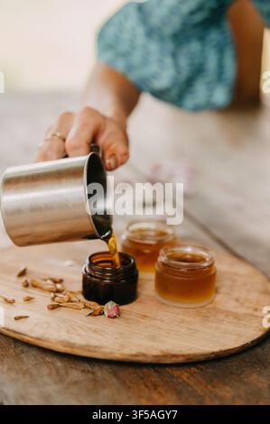 Una mano versa un liquido cosmetico dorato e caldo da una tazza di metallo in piccoli barattoli di vetro su una tavola di legno, mostrando la preparazione di un prodotto di bellezza Foto Stock