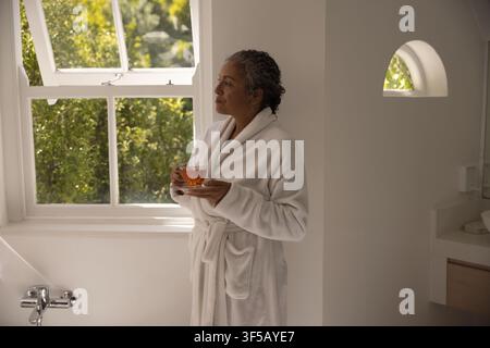 Donna afroamericana di mezza età in veste bianca che tiene una tazza e guarda attraverso la finestra di casa, spazio di copia Foto Stock