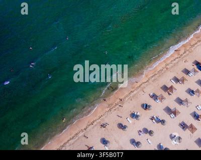 Vista aerea dall'alto con drone di file ordinate di sdraio e ombrelloni di paglia su una spiaggia di sabbia accanto all'acqua turchese chiara del mare sulla costa dell'Ucraina, Foto Stock