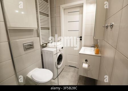 Interno di bagno compatto moderno con piastrelle bianche, lavatrice e caratteristiche minimaliste del WC. Disposizione di piccola lavanderia, design funzionale del bagno, ristrutturazione di appartamento contemporaneo Foto Stock