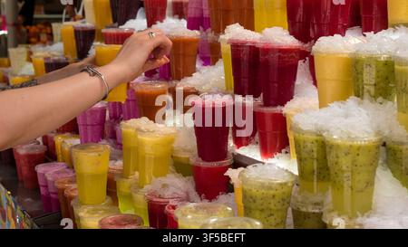 Mano che raggiunge una tazza di frullato di frutta fresca a uno stand di succhi colorato sul mercato su ghiaccio tritato — Stile di vita sano, rinfresco estivo e cibo da strada Conce Foto Stock