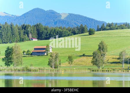 Paesaggio bavarese pittoresco a Bad Bayersoien, Germania, con un lago tranquillo che riflette prati verdi e ondulati, un fienile tradizionale in legno e per Foto Stock