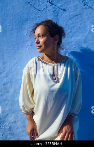 Una donna pensierosa in un abito crema ricamato si trova davanti a un muro blu vivace, illuminata da una calda luce laterale. Ritratto naturale che enfatizza l'atmosfera calma Foto Stock