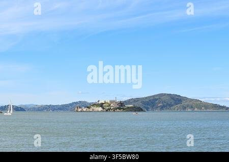 Isola di Alcatraz circondata dalle acque della baia di San Francisco con una barca a vela e le dolci Marin Headlands sotto un cielo blu limpido, California, USA. Foto Stock