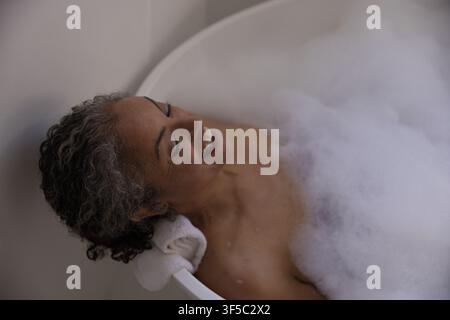 Donna anziana afroamericana sdraiata nella vasca da bagno a casa con schiuma, appoggiando la testa su un asciugamano arrotolato Foto Stock