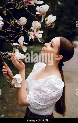 Giovane donna che annusa fiori di magnolia bianchi in un parco verdeggiante in un dolce pomeriggio di primavera alla luce del giorno. Aroma mattutino botanico delicato, serenità femminile nel benessere, parco primaverile in fiore Foto Stock