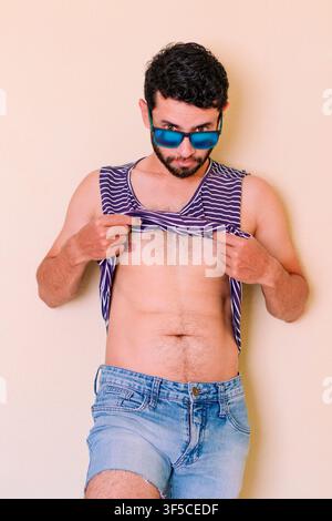 Giovane uomo con occhiali da sole solleva la maglietta, sente il caldo. Uomo in abbigliamento estivo, pescatore con cappello di paglia. Giovane uomo in blu Foto Stock