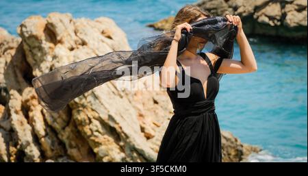Giovane donna in abito nero con sciarpa nera in piedi sulle rocce Foto Stock