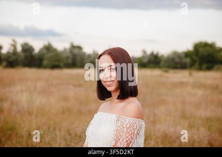 Giovane donna dai capelli scuri in abito bianco di pizzo scollato che posa in un prato dorato al tramonto, catturando la bellezza naturale e la serenità estiva, lifestyle, moda estiva, libertà, natura Foto Stock