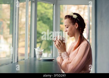 Vista laterale di una giovane donna con un'acconciatura biondo-zingaro intrecciata che tiene una tazza di caffè calda mentre è seduta vicino a una finestra in un ambiente accogliente e tranquillo, atmosfera mattutina confortevole, tempo libero calmo, lifestyle Foto Stock